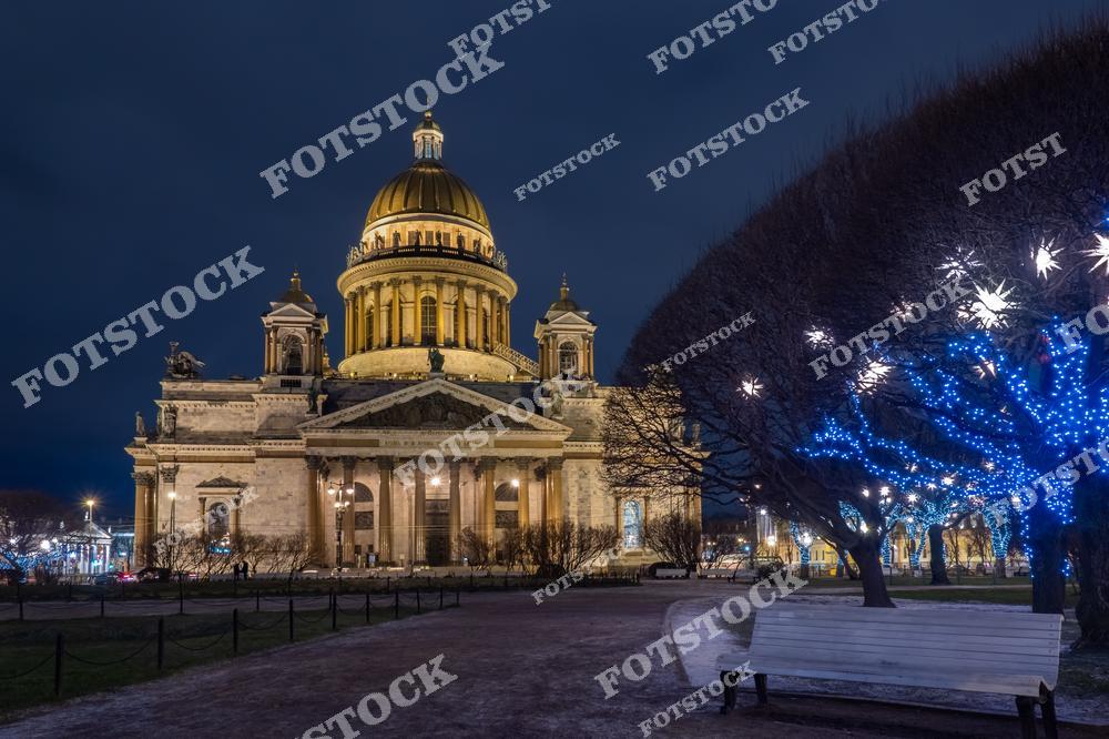 На фотографии изображен Исаакиевский собор, освещенный в ночное время. Его золотой купол и массивная колоннада подчёркнуты мягким искусственным светом. Деревья на переднем плане украшены светодиодными гирляндами, добавляя праздничного настроения. Это место сочетает в себе спокойствие парка и архитектурное величие собора.